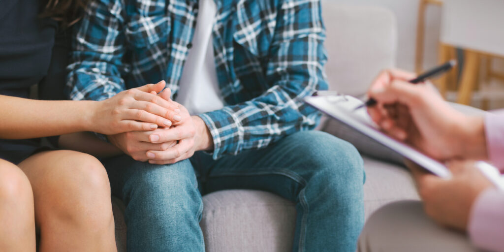 A couple sits together during a counseling session, holding hands as a therapist takes notes in the background. The couples body language suggests they are seeking support and comfort from each other.