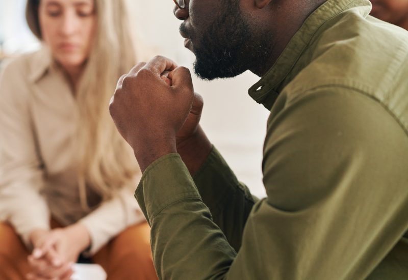 Side view of young stressed man with interlocked hands by his face sharing his problem while attending psychological session