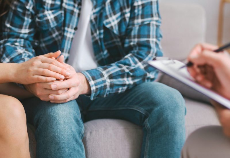 A couple sits together during a counseling session, holding hands as a therapist takes notes in the background. The couples body language suggests they are seeking support and comfort from each other.